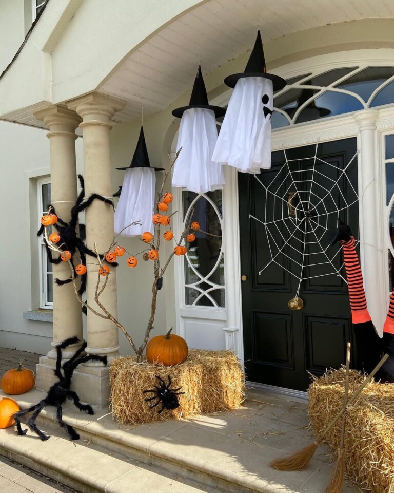 Front entry decorated with hay bales, giant spiders, pumpkin lights, ghost figures in witch hats, and a large spider web on the door.