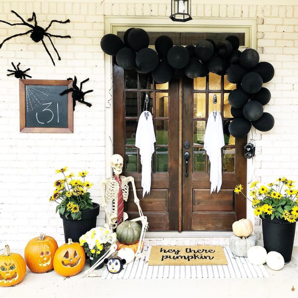 Wooden double doors with black balloon garland, skeleton figure, ghost cutouts, pumpkins, and yellow flowers in planters.