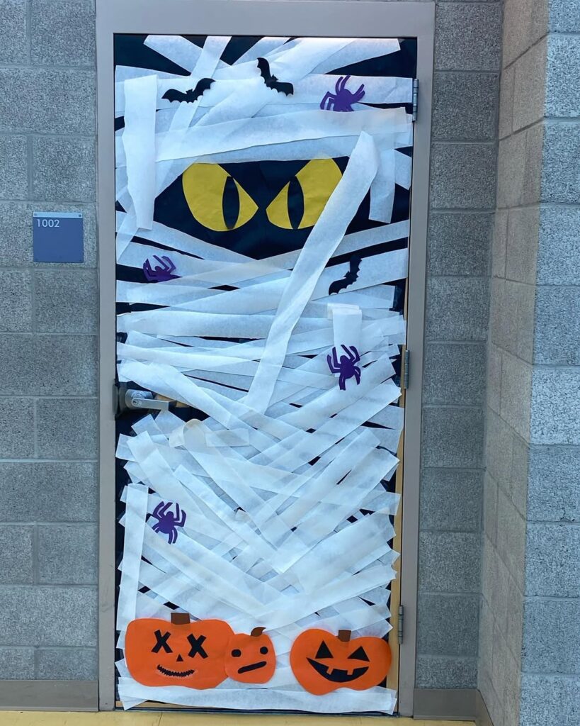 Mummy-themed classroom door wrapped in white streamers with glowing yellow eyes, bats, spiders, and pumpkin cutouts at the bottom.