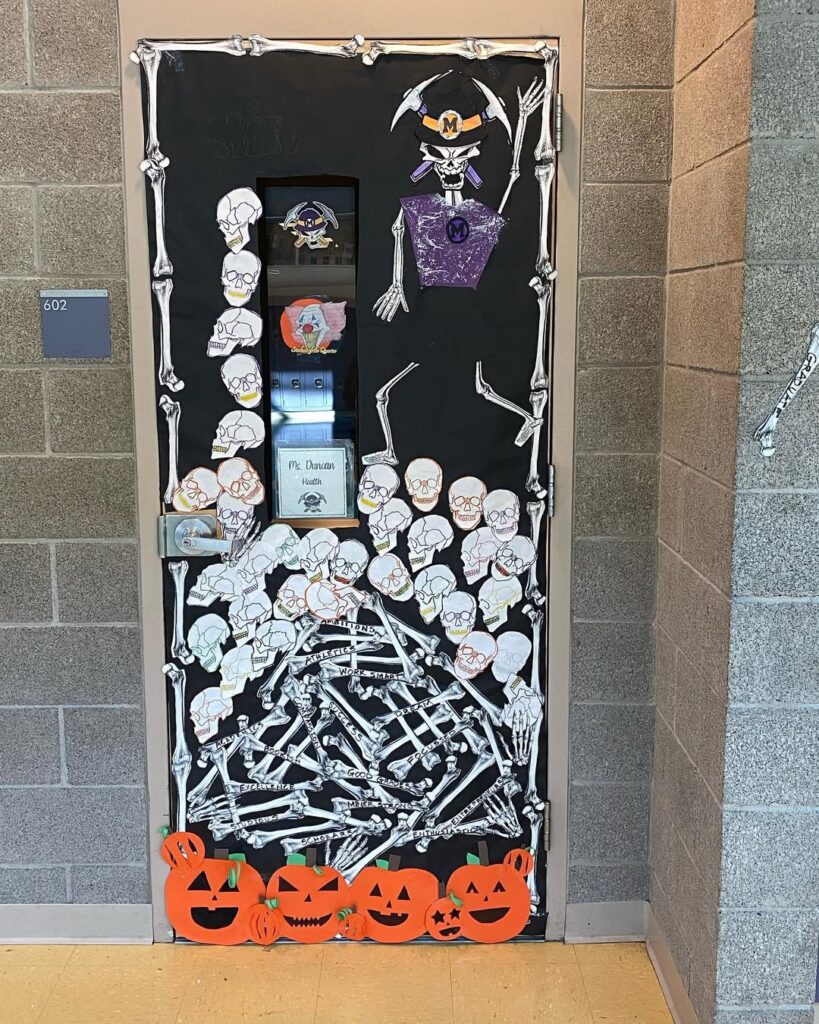 Classroom door decorated with skeleton cutouts, skull drawings, orange jack-o’-lantern cutouts, and black backdrop.