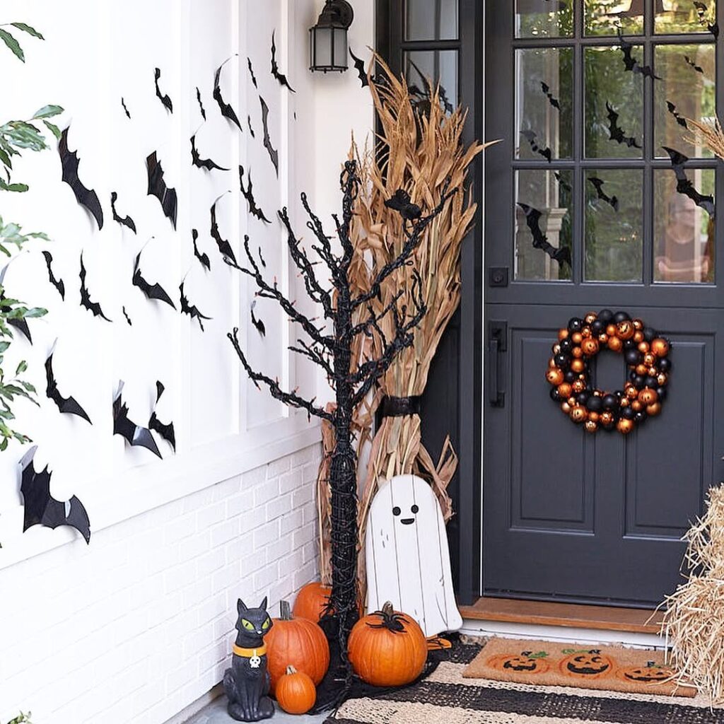 Halloween porch with flying bat cutouts, pumpkins, a ghost decoration, corn stalks, and an orange-and-black wreath on the door.