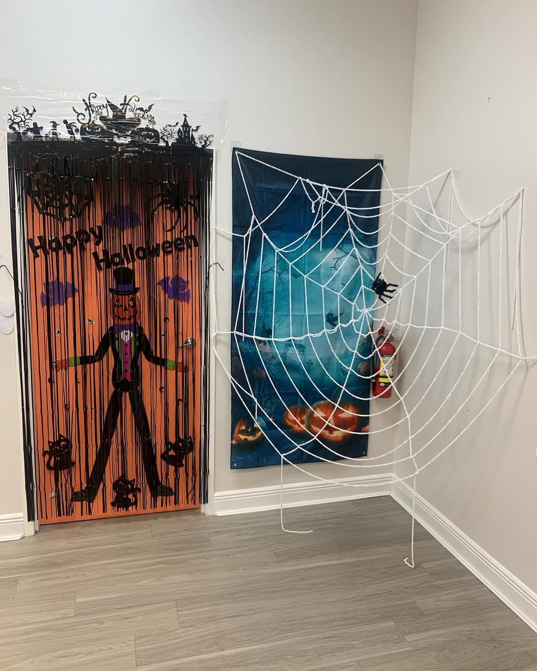 Indoor Halloween display with giant white spider web, pumpkin-headed figure, bats, black cats, and an orange "Happy Halloween" door backdrop.