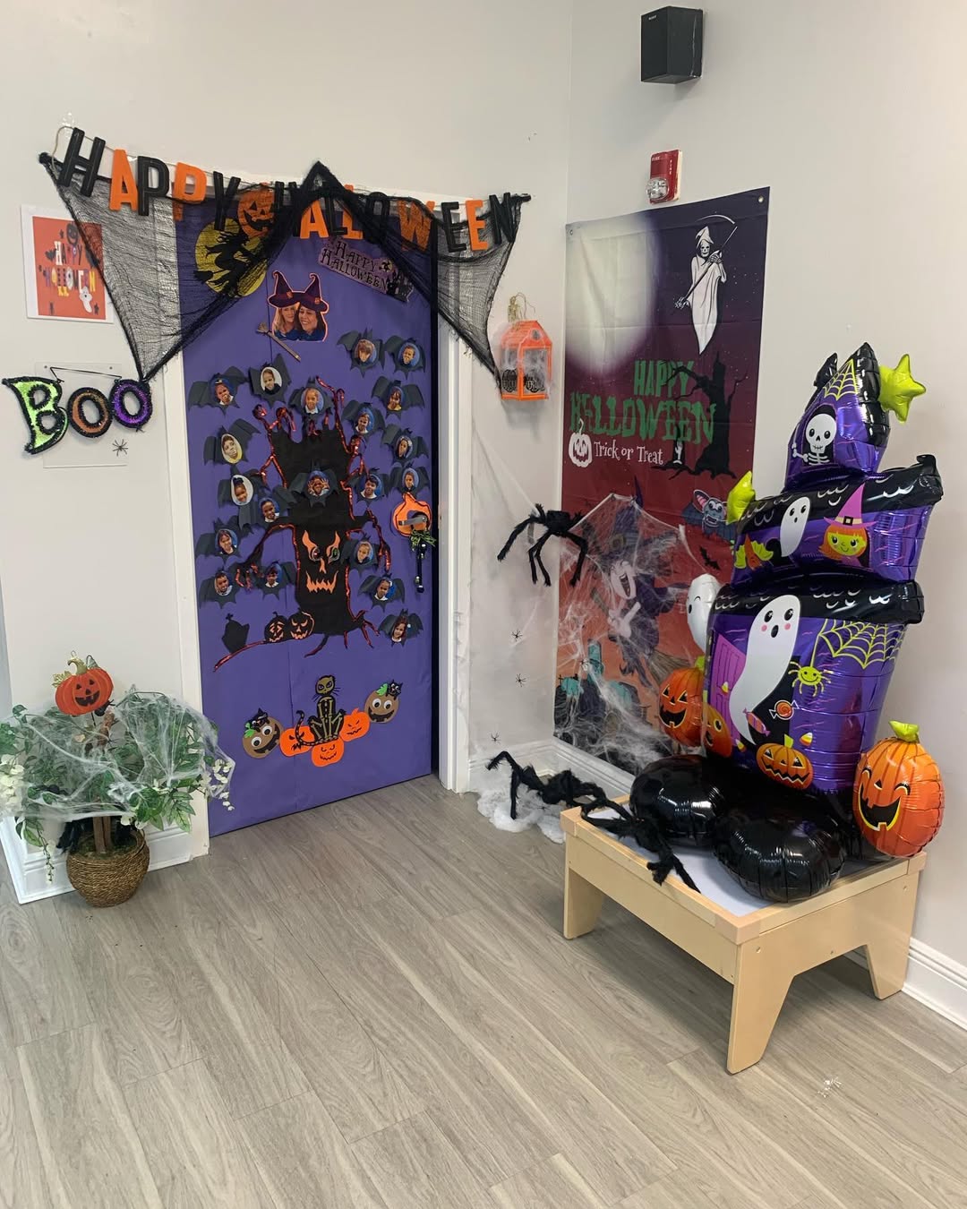 Halloween classroom door decorated with purple backdrop, cartoon faces, bats, pumpkins, and a "Happy Halloween" banner, surrounded by spiders and ghost inflatables.