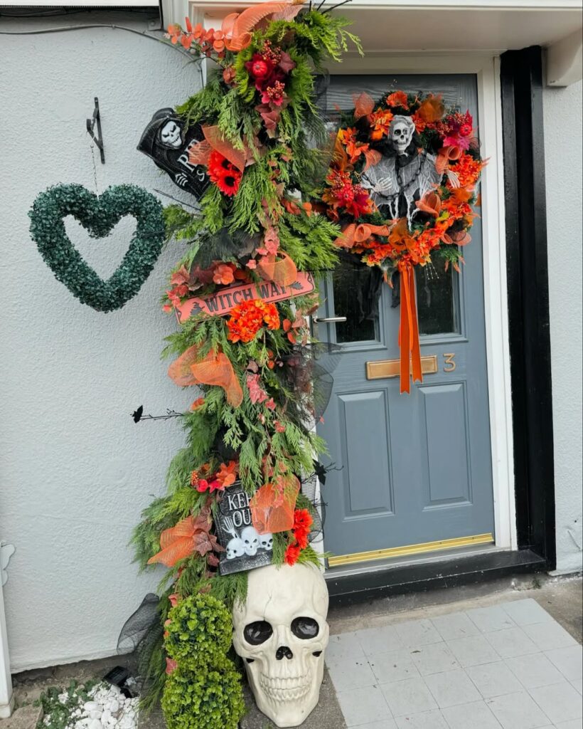 Grey front door with orange-and-black Halloween wreath, greenery garland decorated with skulls and skeleton signs, and large skull at the base.