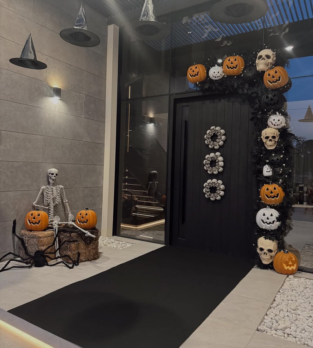 Modern black front door framed with pumpkins, skulls, and skeleton wreaths, with floating witch hats and a skeleton sitting by hay bales.