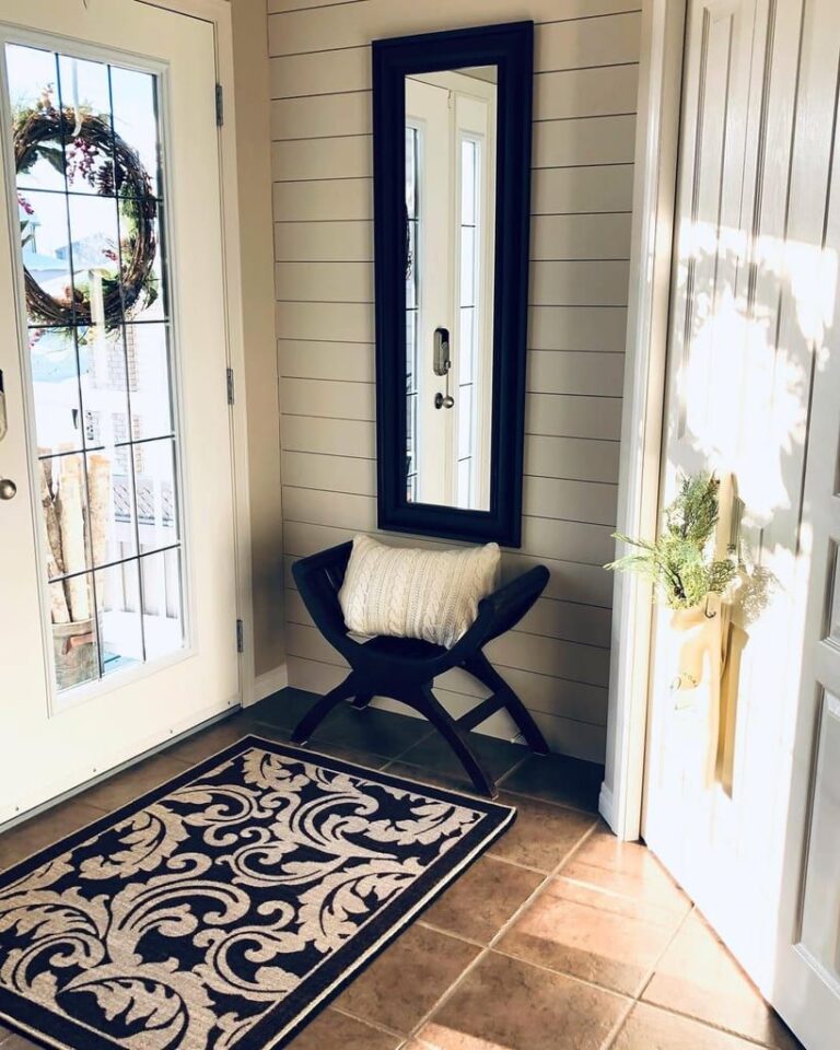 Small entryway decorated with a large dark framed mirror, patterned black and cream rug, and light shiplap walls.